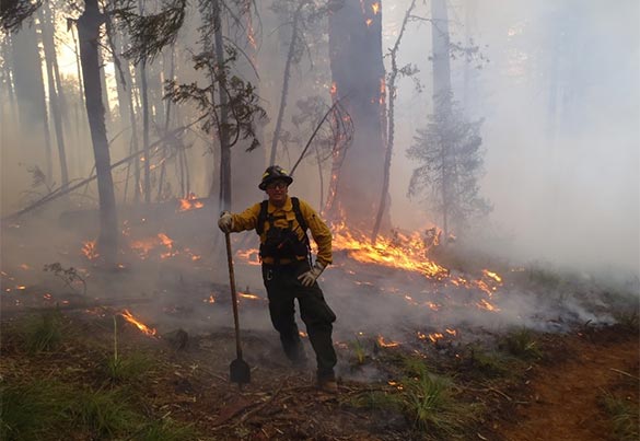 A man wearing fire gear leans on his shovel in front of a low intensity fire.