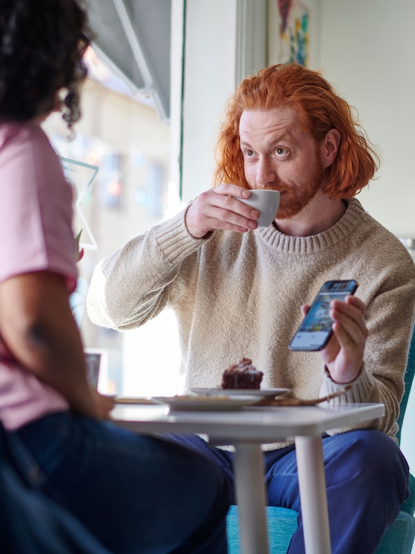 Un hombre y una mujer sentados a la mesa de un restaurante IKEA mientras mantienen una conversación. El hombre está bebiendo de una taza y sostiene un móvil.