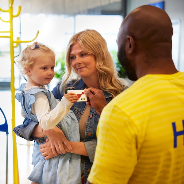 Una mujer sostiene en brazos a una niña pequeña mientras recibe una tarjeta regalo IKEA de un colaborador con una camiseta amarilla, en una tienda IKEA.