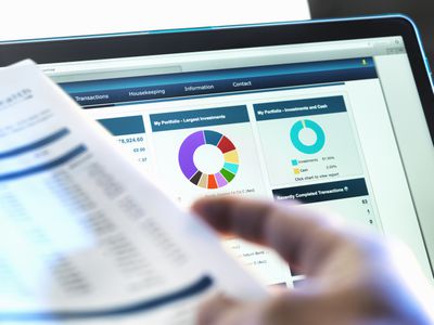 A computer screen close up showing colorful charts for an investment porfolio, while an out of focus hand in the foreground holds paperwork