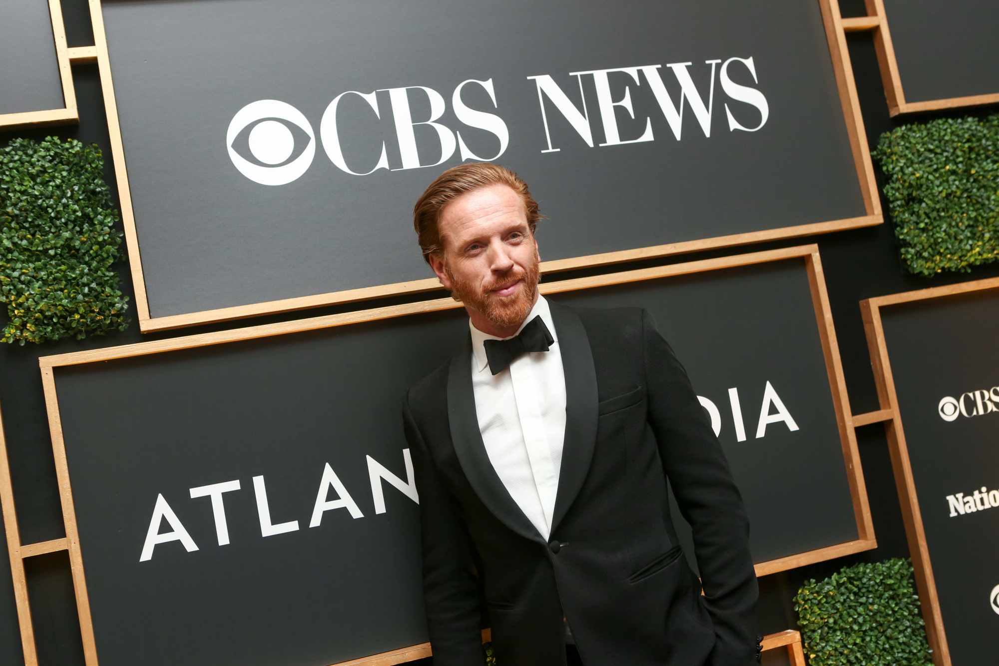 Billions actor Damian Lewis poses in a tuxedo under a CBS News logo