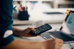 A person takes notes while looking at financial charts on a smart phone