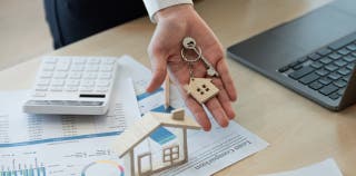 Woman's hand holding a key with a house-shaped keychain while loan papers, a calculator, and a laptop are in the background