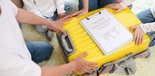 Four people kneel down, each with their hands on the same suitcase which has a travel insurance policy resting on it.