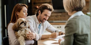 A man and woman sit at a table, signing pet insurance documents. The woman is holding a shih-tzu in her arms.