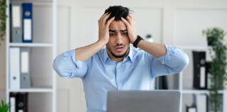 A distressed young businessman sits at his desk with a laptop open in front of him