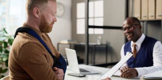 A man with his arm in a sling is looking at a workers' compensation document while another man helps explain it to him 