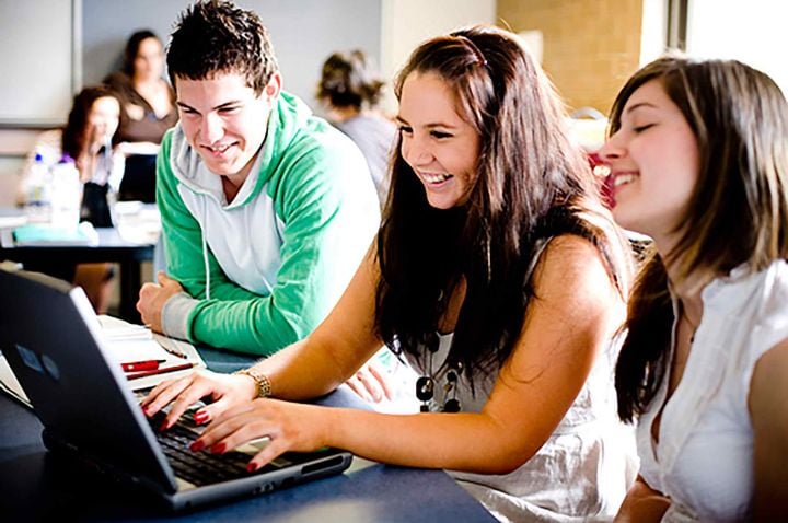 male and two female students in a classroom, smiling and using a laptop