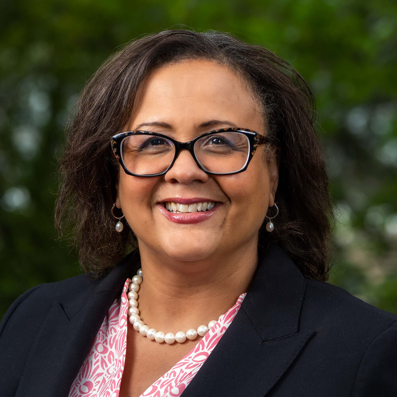 Dark-haired person with glasses wearing a pink shirt, pearls, and a black blazer.