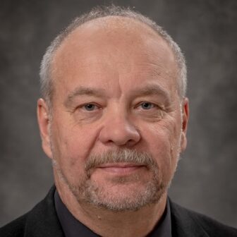 Robert Kostecki, a short-haired person poses for a headshot against a gray background.