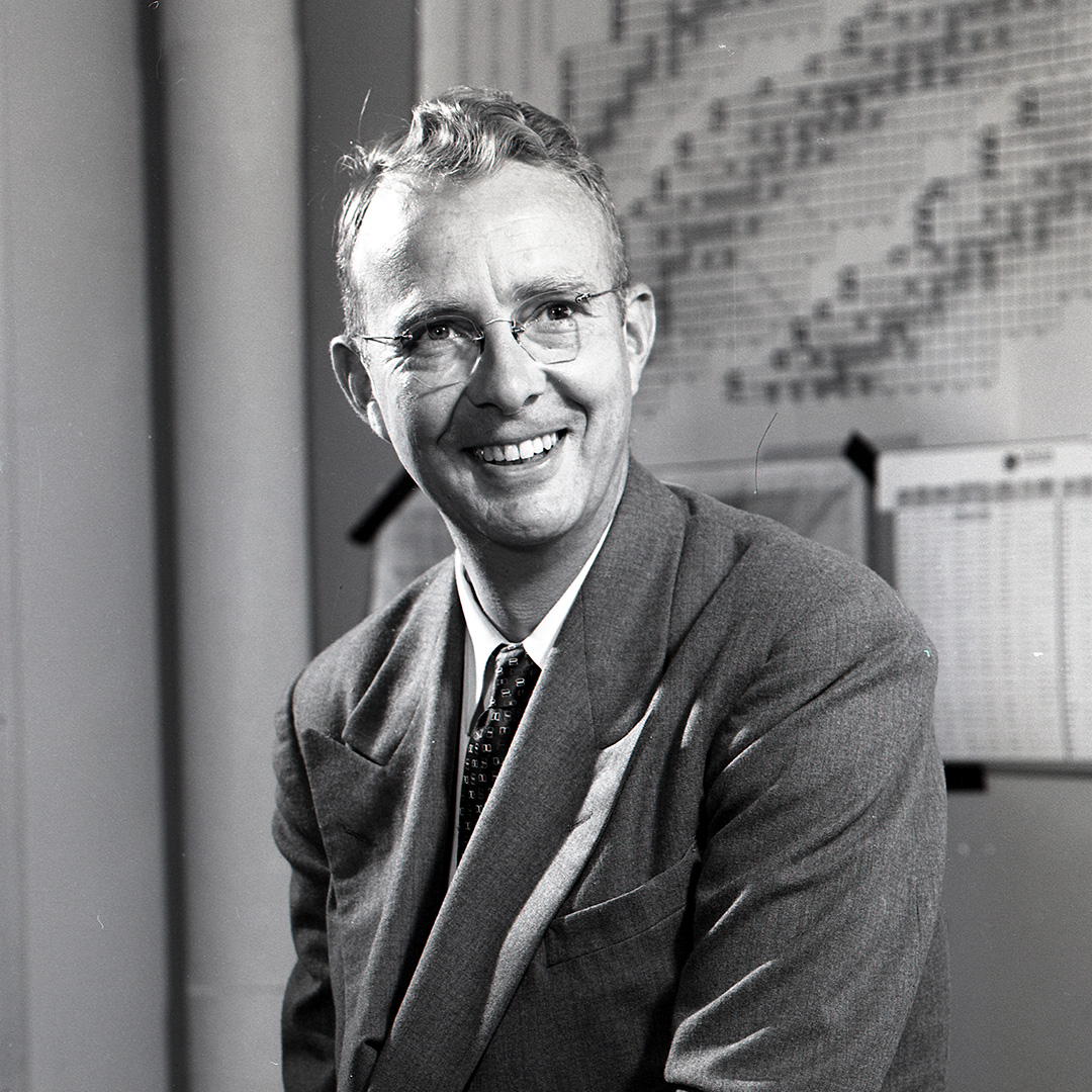 Black and white portrait of Luis Alvarez, a short-haired person smiling in a suit and tie.