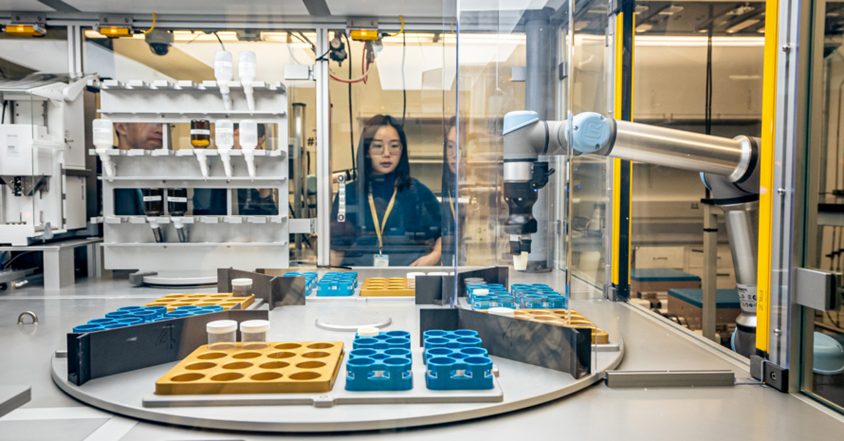 Dark-haired scientist in the center of the frame looks toward the camera. They are standing behind the clear glass of an encased automated lab.