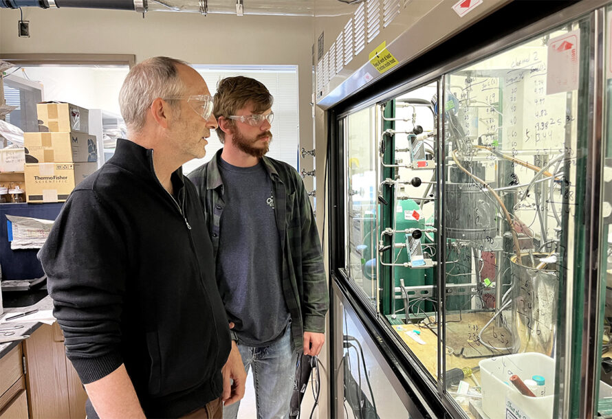 Two people in safety glasses observe a scientific experiment through a glass window in a lab, with equipment and handwritten notes visible inside the fume hood.