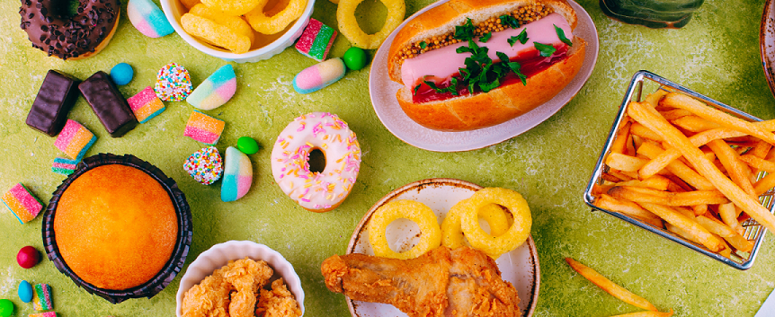 Viele ungesunde Speisen wie Burger, Pommes und Donuts stehen auf einer hellgrünen Oberfläche.