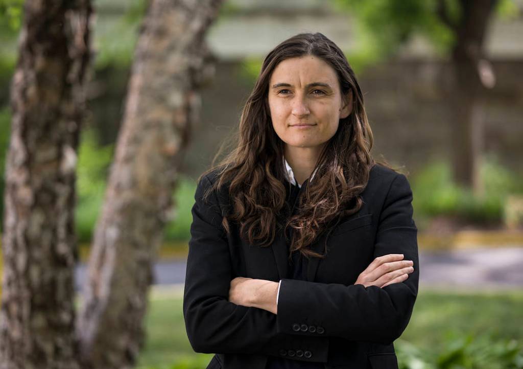 A woman with dark, long brown, wavy hair looks into the distance with a soft smile on her face. She's wearing a black blazer and the collar of her white button down shirt sticks out the top. A tree and greenery is blurred in the background.