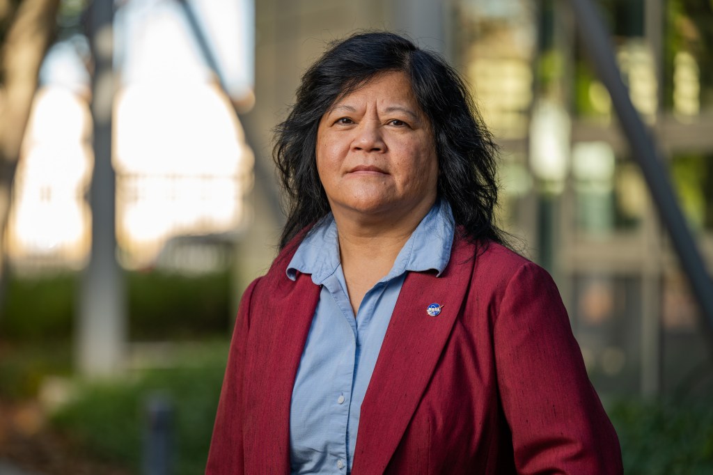 A Vietnamese-American woman with shoulder-length black hair stands in front of a building that is out of focus. She wears a light blue shirt with buttons and a red blazer with a small NASA "meatball" pin.