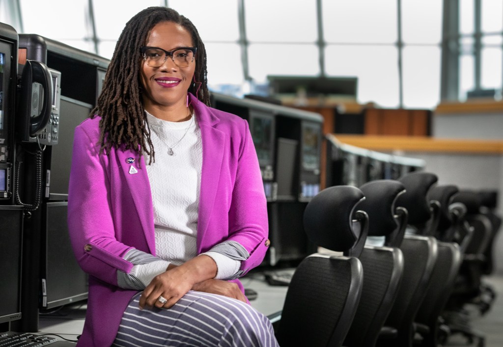 Ales-Cia Winsley smiles for the camera as she sits in front of a row of desks in Firing Room 1 in the Launch Control Center at NASA's Kennedy Space Center. She is wearing a bright magenta jacket with two pins (the NASA "meatball" insignia and the NASA Artemis logo) present on her right lapel, a white shirt, and striped slacks.