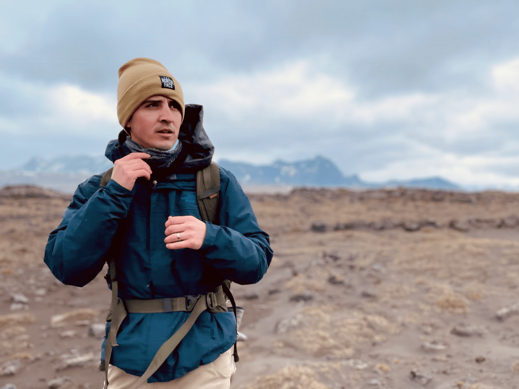 A man wearing a tan beanie and a blue winter jacket with a hiking backpack strapped around him. He's walking across a rocky surface with mountains in the background.