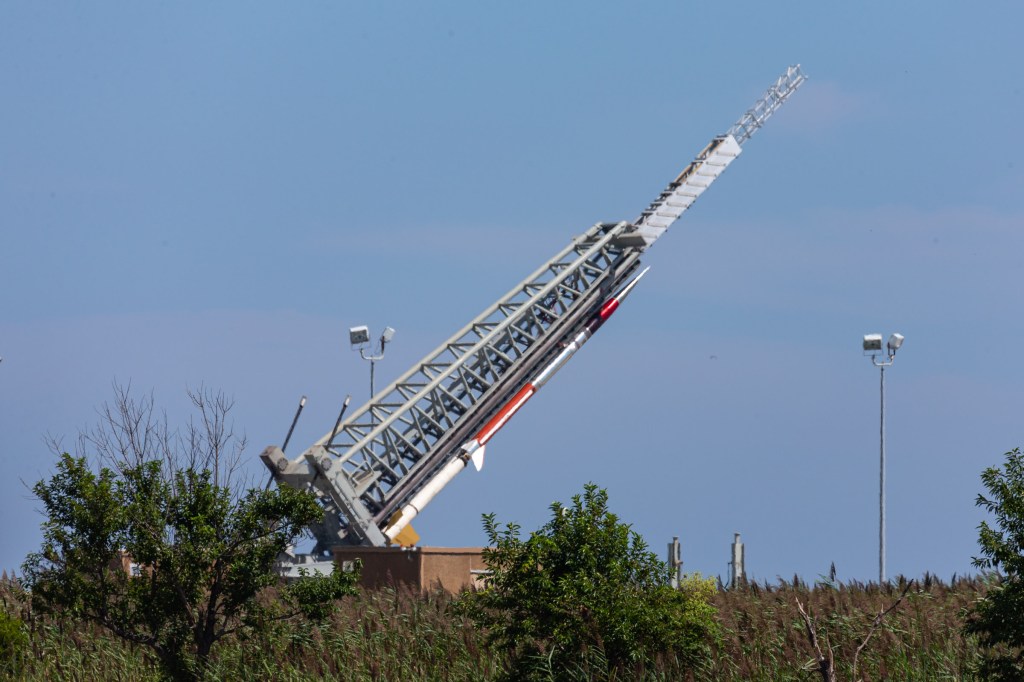 A sounding rocket is being raised on the launch rail, it is still in a horizontal position. The red a silver rocket contrasts against the bright blue sky behind, and green foliage can be seen at the bottom of the photo