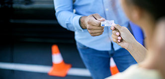 Close-up view of one person handing a driver's license to another person. Two orange traffic cones are on the ground in the background.