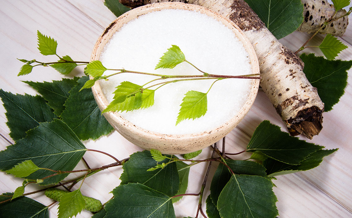 Xylitol crystals in a birch bowl beside a birch branch with leaves.