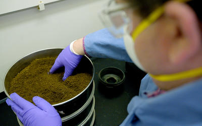 A person wearing a mask and laboratory gloves handles ground plant material inside a metal container.