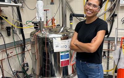 Cassie Stoffer smiles while wearing safety glasses and standing next to a large metal tank with many pipes and wires attached to it. 