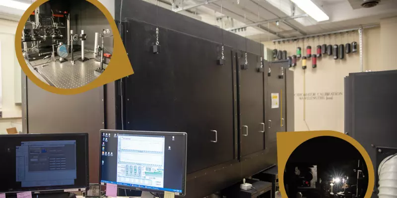 A high-tech laboratory with large black equipment featuring two circular insets showing detailed machinery setups. A computer workstation is in the foreground.