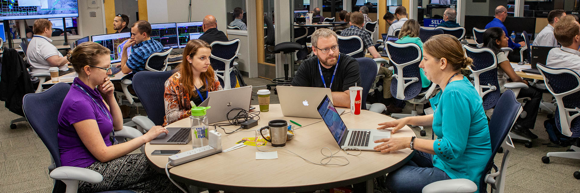 Three women and one man working with laptops at a round table in a room full of many workers in front of computers and large dispslays