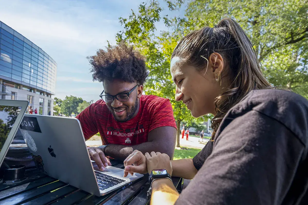 A man wearing glasses and a red shirt and a woman with dark hair wearing a black t-shirt sit looking at a laptop and smile