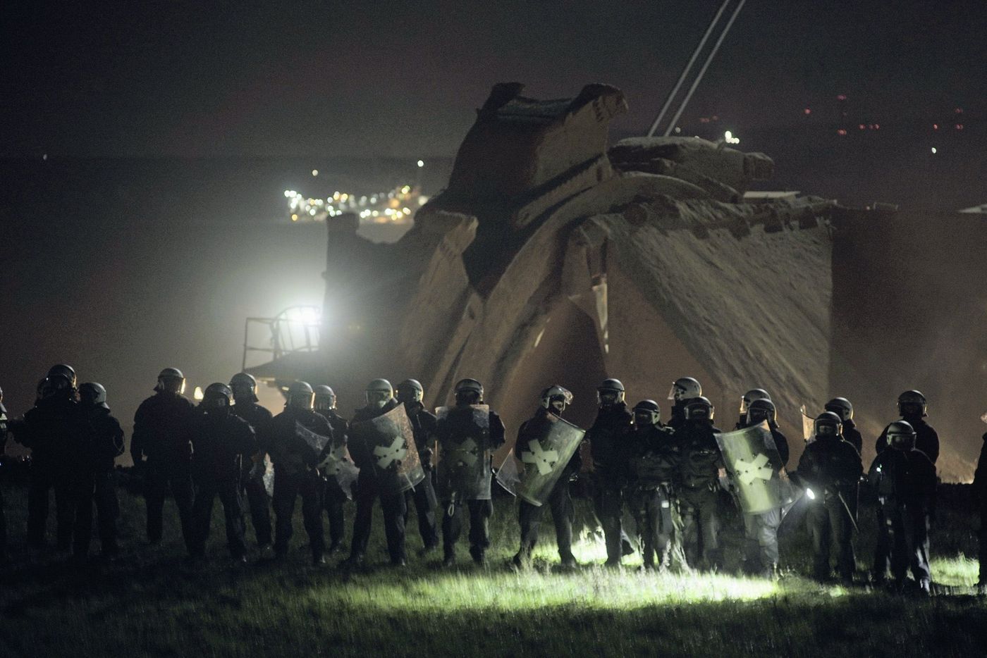 "A spookily lit picture of a group of riot cops, helms, shields and all, standing in front of a gigantic bucket excavator."