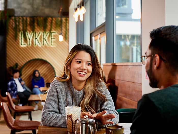 Students in a cafe in Sheffield city centre. A young woman smiles at a young man across the table from her. There is a milkshake glass and a stainless steel pot on the table. In the background we can see a spacious room lit by hanging bulbs and floor-to-ceiling windows, with wooden chairs, benches and wall panels.