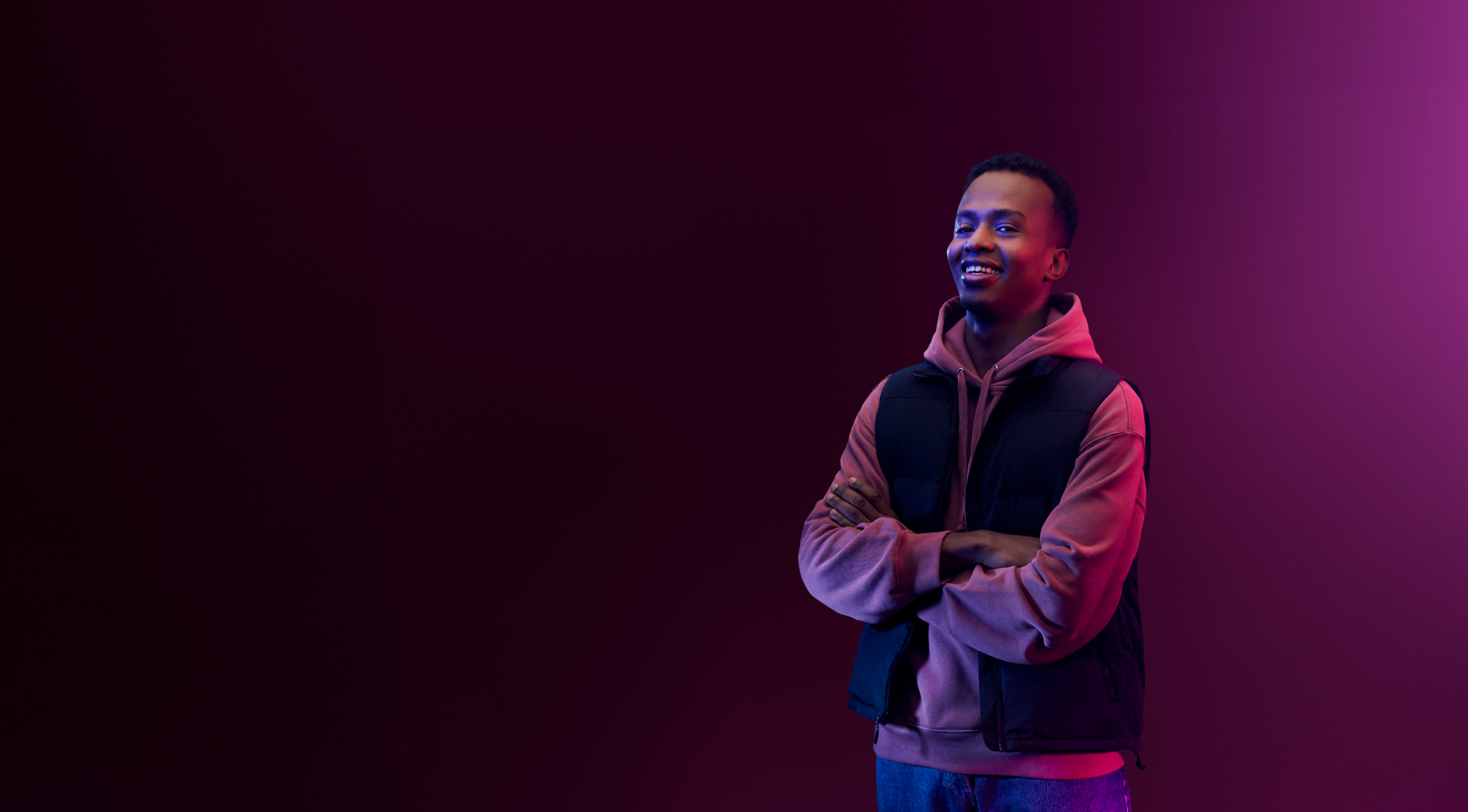 Purple background with student in a body warmer over a hoody smiling with their arms folded