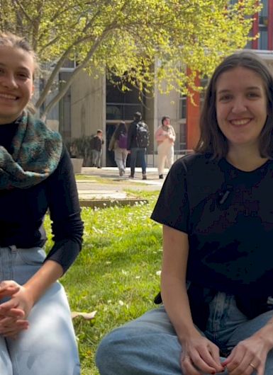 Two young women sitting on the grass at a university campus, smiling.