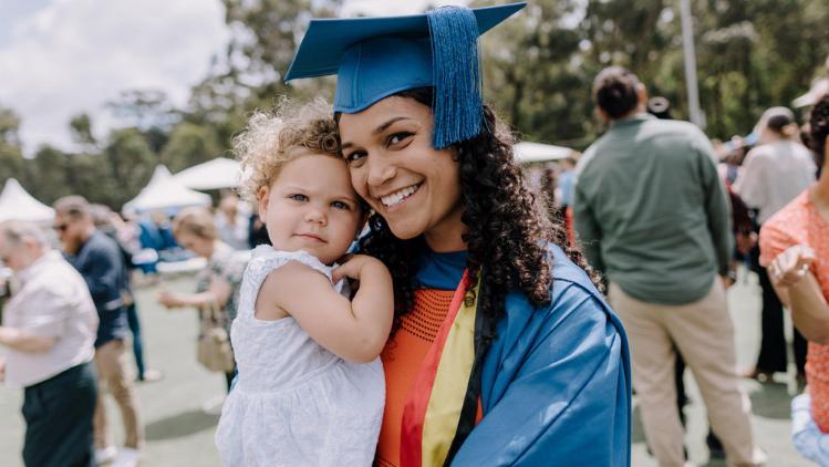 A recent graduate stand on the Uni Active soccer fields with her daughter on her hip. Both are smiling at camera, after the graduation