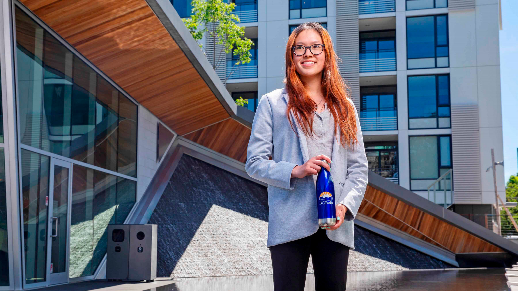 A student with long hair stands in front of a building holding a BC Parks water bottle.