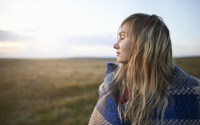 A woman stands with her eyes closed and wrapped in a blanket on a moorland.