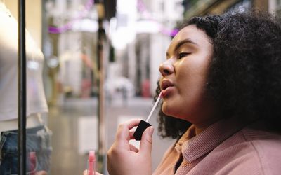 Young woman with curly hair applying lip gloss in front of store window