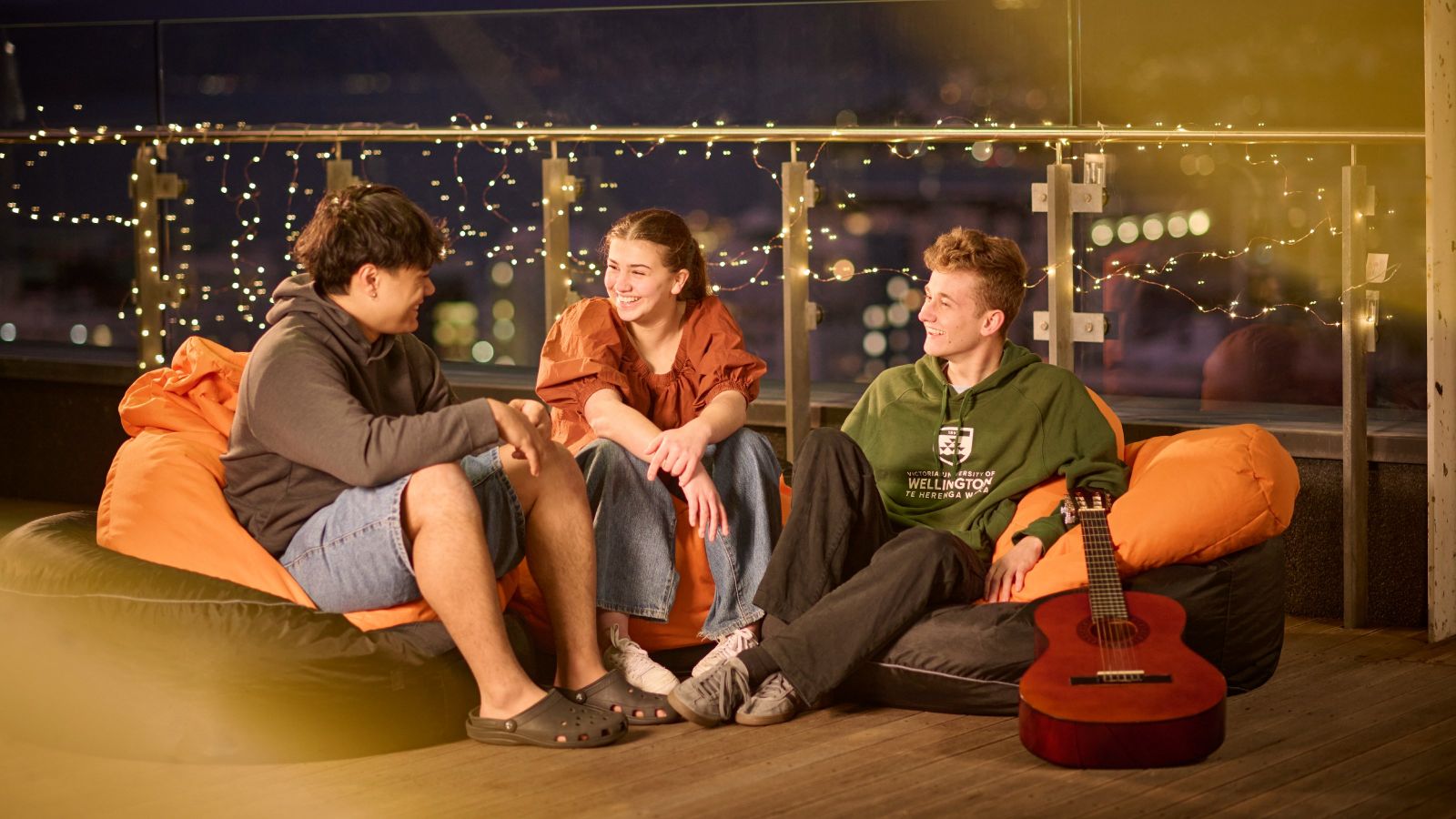 Three residents sitting on beanbags on a balcony at a hall of residence at night, with a guitar