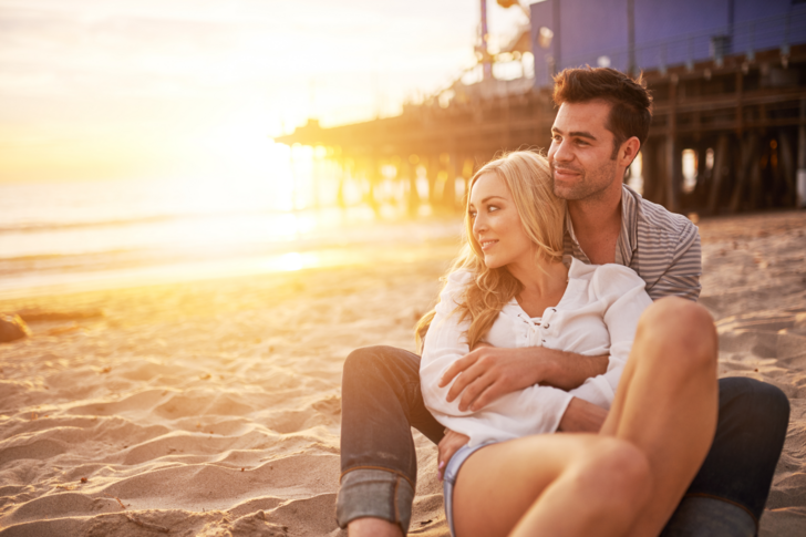 Um homem abraça uma mulher por trás, enquanto assistem, sentados e felizes, o pôr do sol na praia.