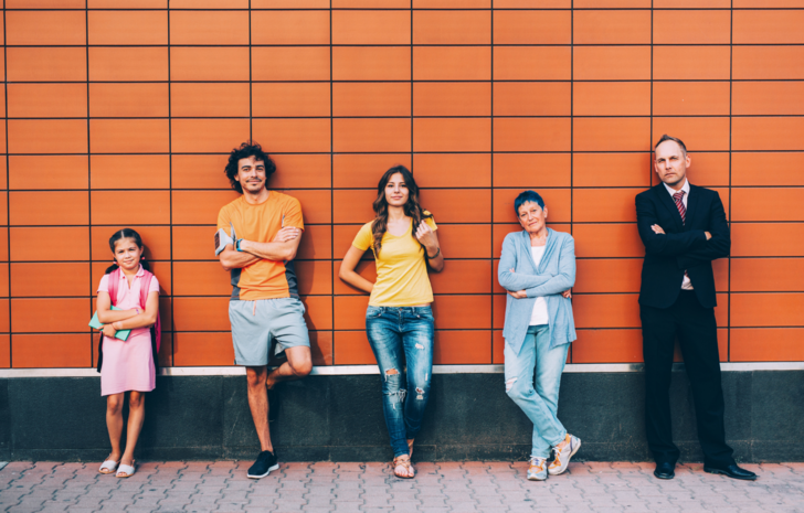 Uma garotinha, dois jovens e dois adultos estão em pé, em fila, apoiados na parede.