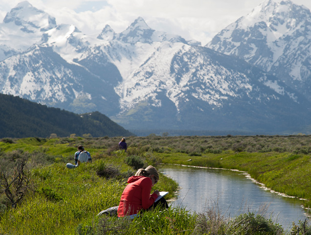 A student sitting near a stream that leads to large mountains in the background